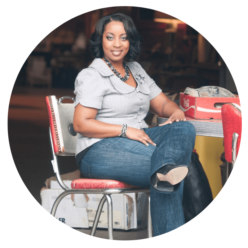 Team Sesi. Headshot of Jeana Aaron, contributing layout designer. She's a Black woman, and in this photo, she's seated in a red and white chair by a table. Her hair is short, one leg is crossed over the other, and she's wearing dark denim jeans, a taupe blouse, and a blue, black, and taupe necklace.
