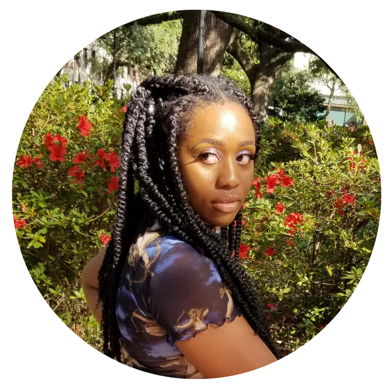 Team Sesi. Photo of Ryan Oakley, contributing illustrator. She's wearing large Senegalese twists and is standing outside by a flower bush. She's looking off to one side and is wearing a blue and black short-sleeved top with angels on it.
