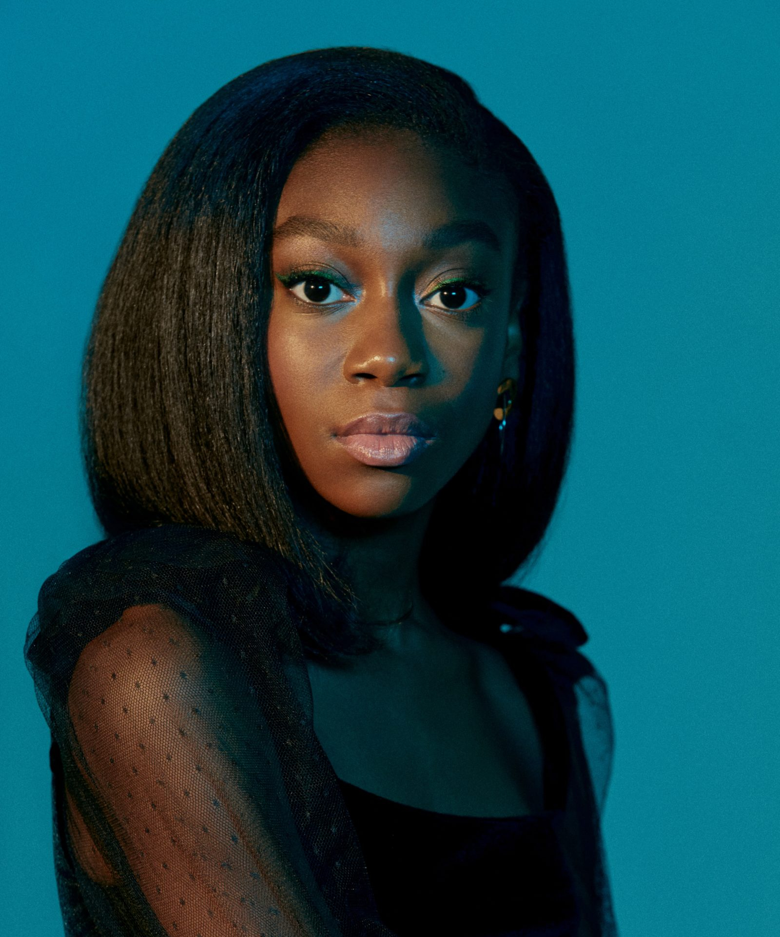 Shahadi Wright Joseph wearing black top with sheer sleeves against blue background