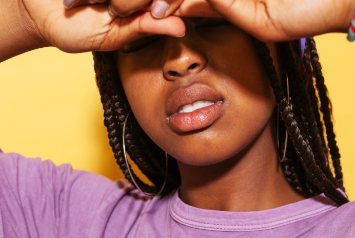 Stylish African-American teen girl in violet outfit pulling finger in front of closed eyes against yellow background. Her hair is in braids and she's wearing gold hoop earrings and a purple head scarf.