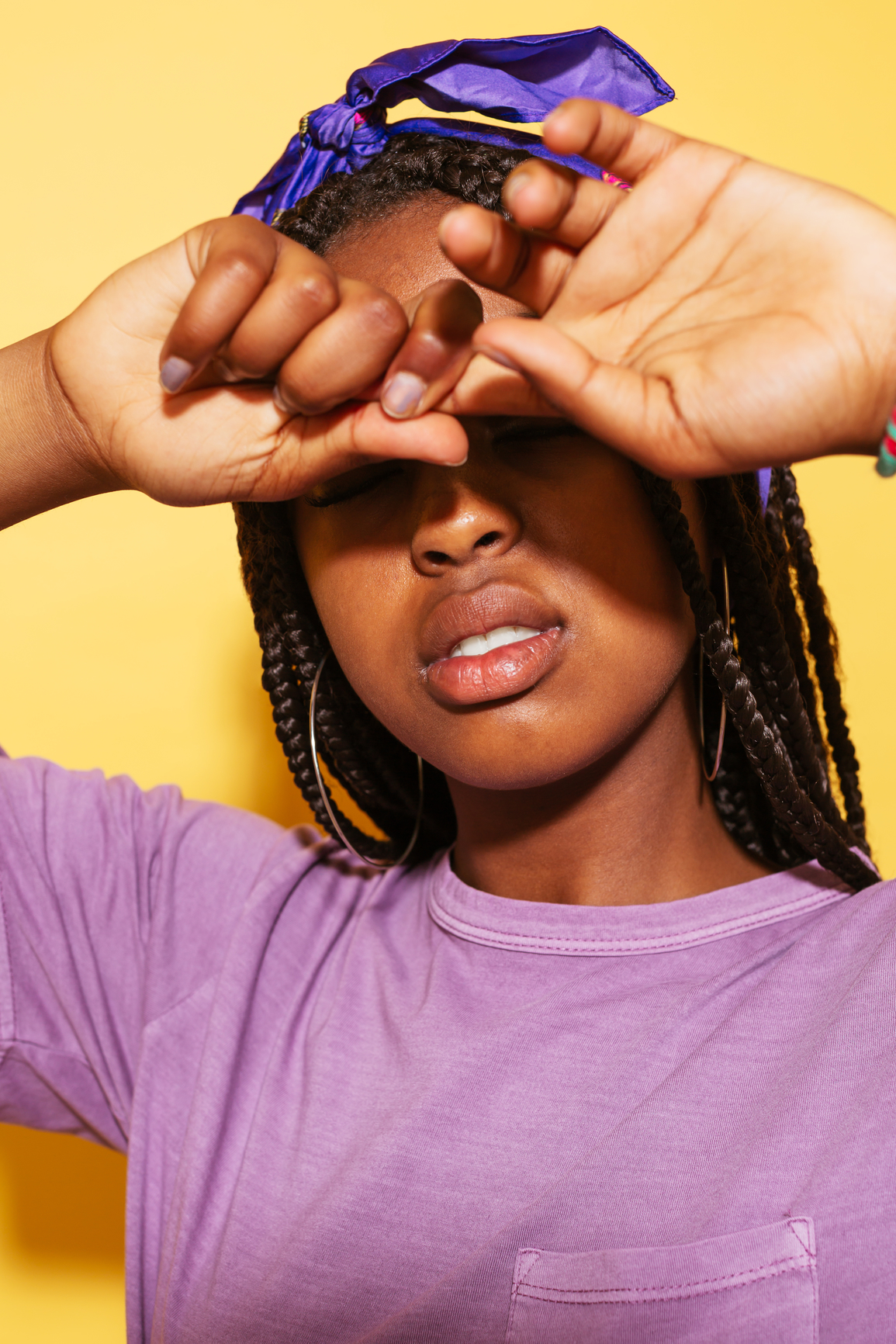 Stylish African-American teen girl in violet outfit pulling finger in front of closed eyes against yellow background. Her hair is in braids and she's wearing gold hoop earrings and a purple head scarf.