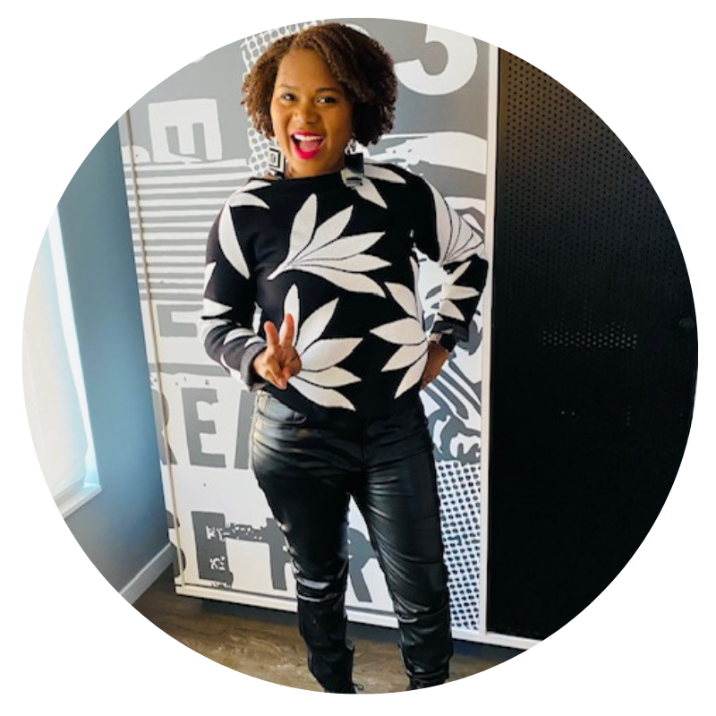 Team Sesi. Photo of Laswhawn Hudson, contributing writer. She's wearing a curly afro, a black sweater with white flowers/leaves and black leather pants. She's smiling and posing with one hand on her hips and one making the peace sign.