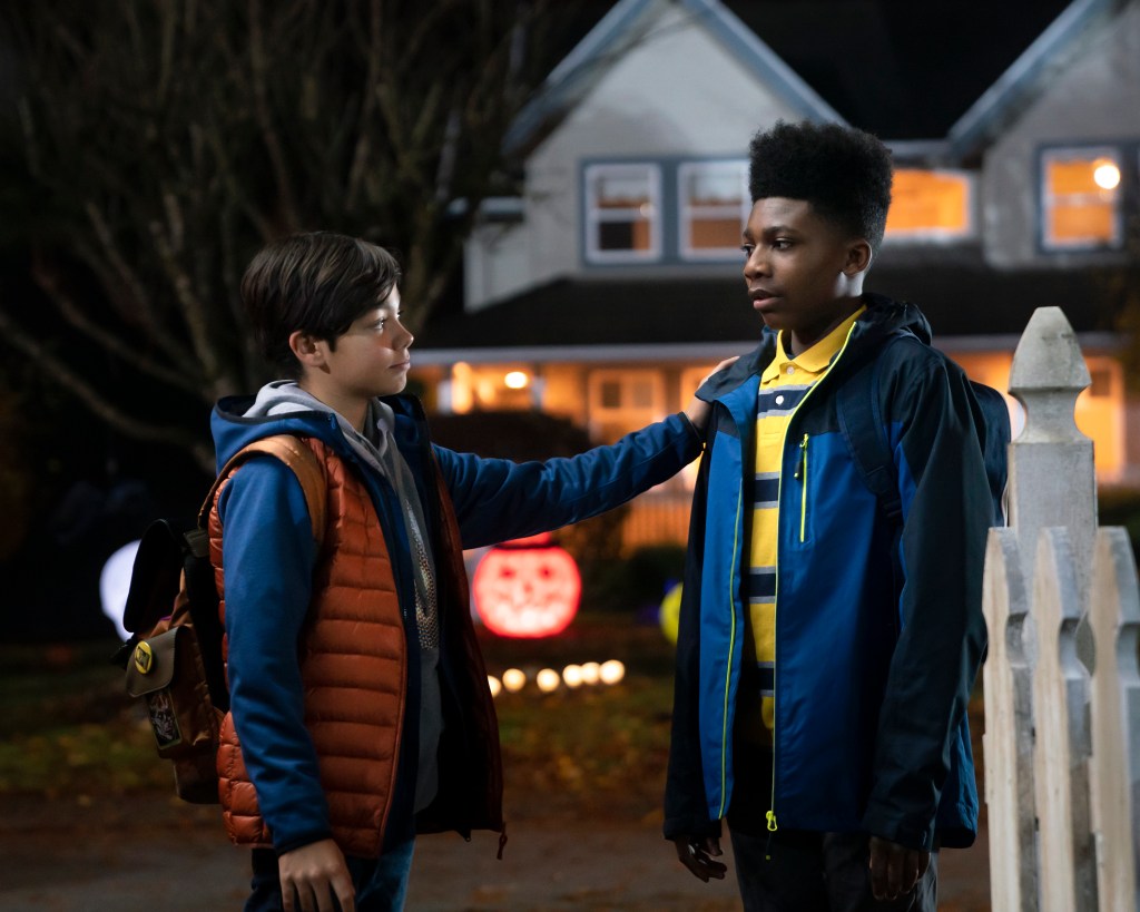 Christian J Simon, wearing a yellow and blue striped shirt, dark blue jacket, and backpack stands next to co-star Malachi Barton in front of a house decorated for Halloween in this still from "Under Wraps."