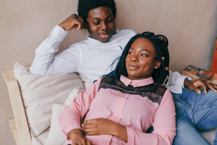 a young, Black teen girl is resting on the lap of her boyfriend. Both have light smiles on their faces.