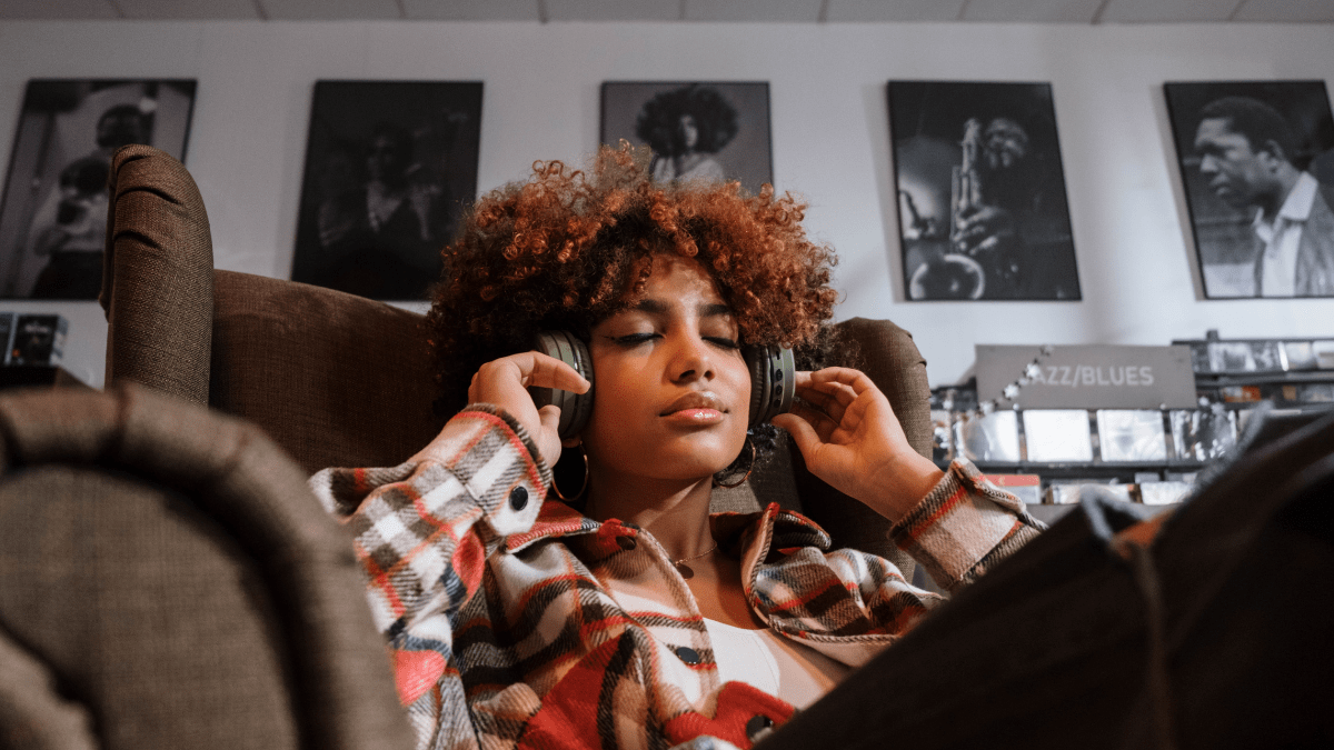 Black teen girl with curly afro listening to music on headphones while sitting in a chair in a record shop