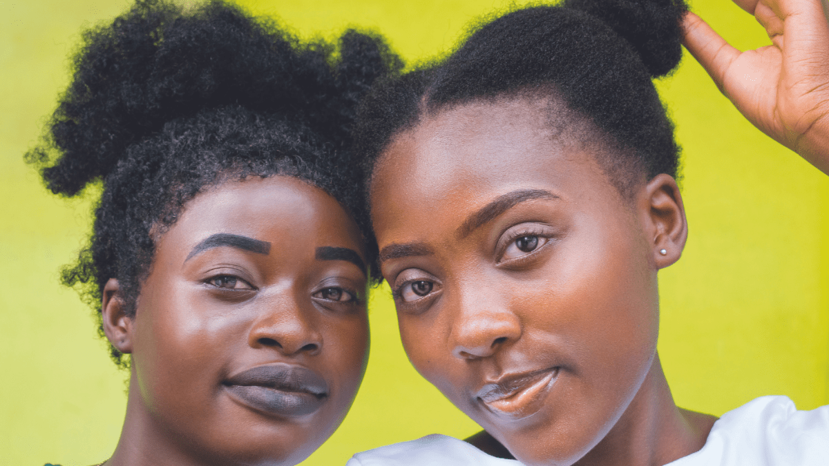 Two dark-skinned Black teen girls with natural hair facing the camera; one has hair in a curly top pony and another in two buns