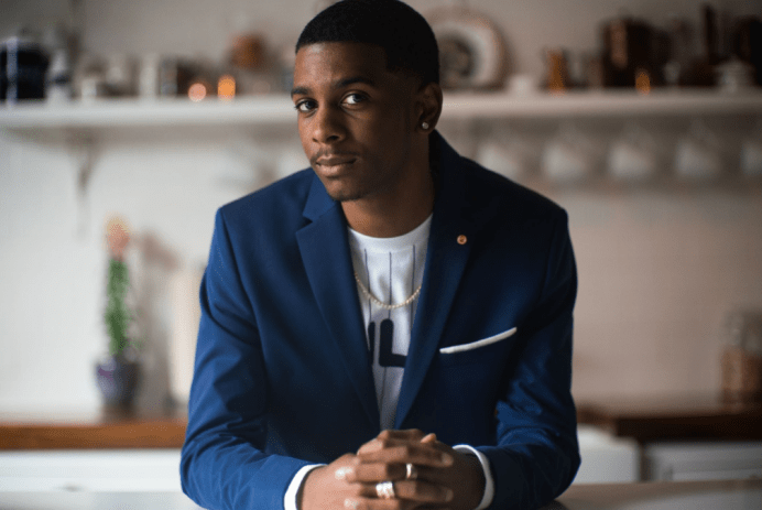 Brett Gray wearing a dark blue blazer over a white tee while sitting at a table with hands folded in front of him