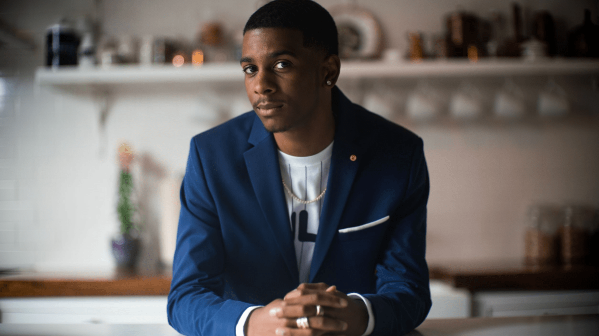 Brett Gray wearing a dark blue blazer over a white tee while sitting at a table with hands folded in front of him