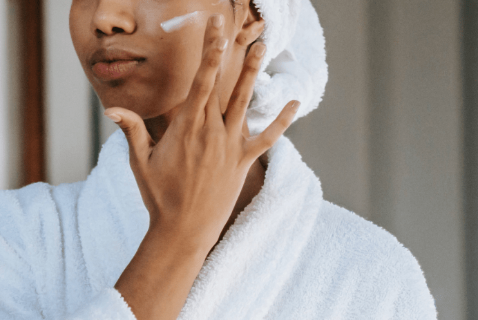 Black teen girl applying face cream while wearing white robe and white towel on head