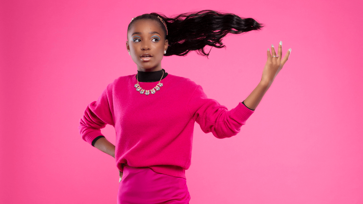 Saniyya Sidney on pink background wearing pink sweater and pink skirt. Her ponytail is captured floating mid-air to the side