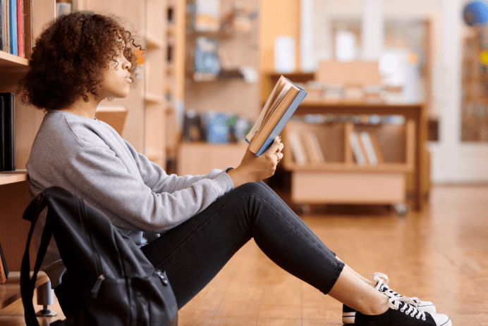 Main image for article about the GOP banning books. It depicts a Black teen girl with curly hair sitting on the wood floor in a library reading a book. She's wearing a gray sweatshirt, dark jeans, and black and white tennis shoes. A black backpack is on the floor next to her.