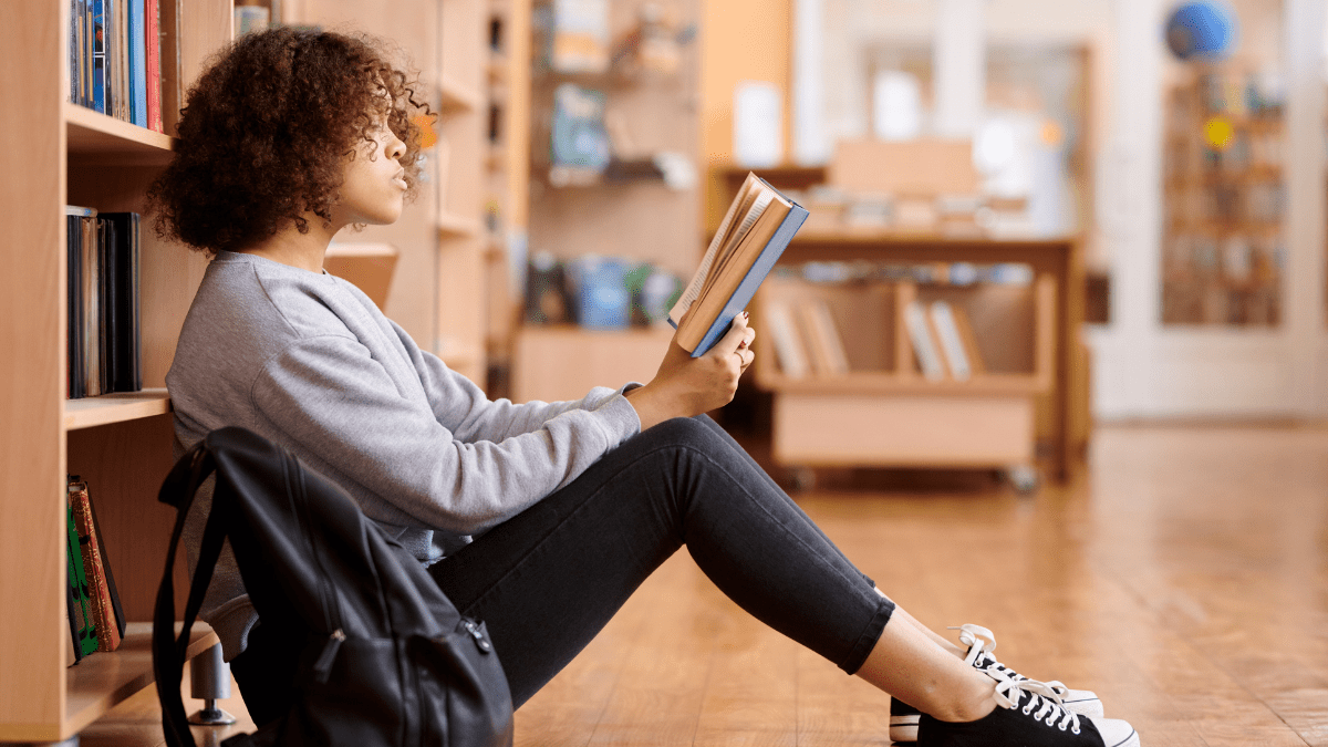 Main image for article about the GOP banning books. It depicts a Black teen girl with curly hair sitting on the wood floor in a library reading a book. She's wearing a gray sweatshirt, dark jeans, and black and white tennis shoes. A black backpack is on the floor next to her.