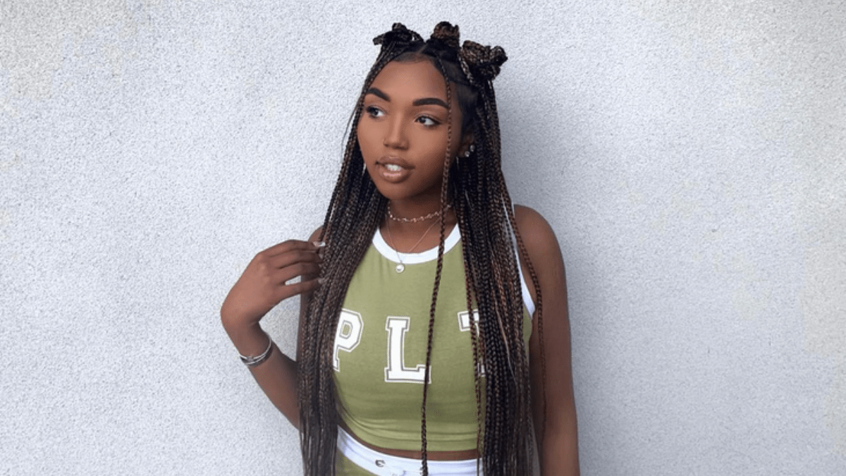 Sy'rai standing against a gray wall wearing a green and white tank top and joggers. Her hair is in long braids in a half-up of Bantu knots and half-down style